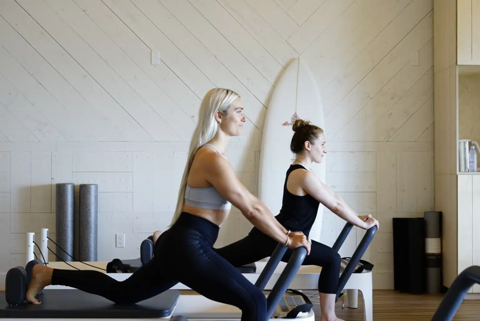 Two women in athletic wear are working out in Montauk, New York Pilates style on reformer machines in a well-lit studio with wooden walls and a surfboard propped in the background, both focused and stretching their back legs.
