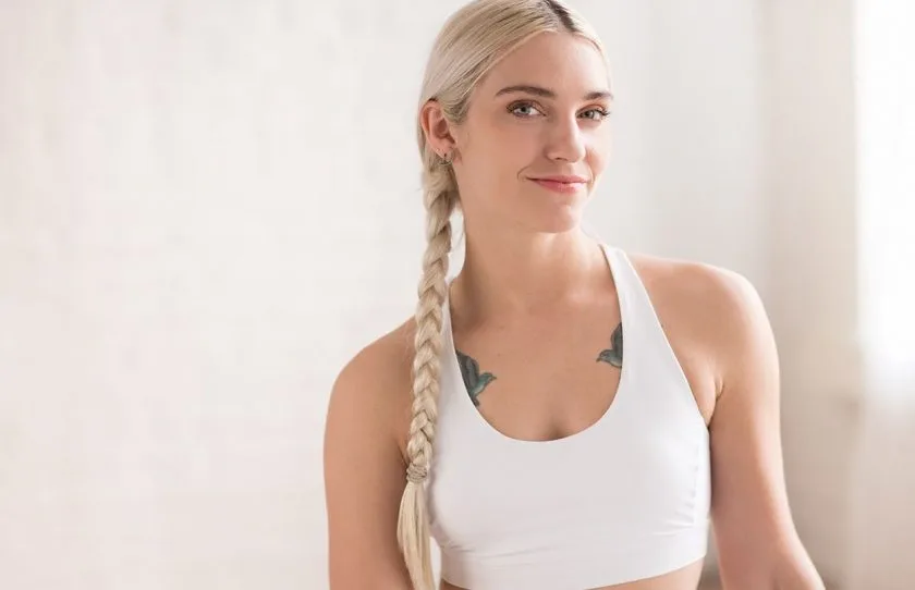 A woman with long blonde hair in a braid, wearing a white sports bra, smiles slightly while standing in a bright, softly lit room. She has visible tattoos on her chest—ready for her NEW YEAR’S RESOLUTION RESCUE at NEW YORK PILATES.