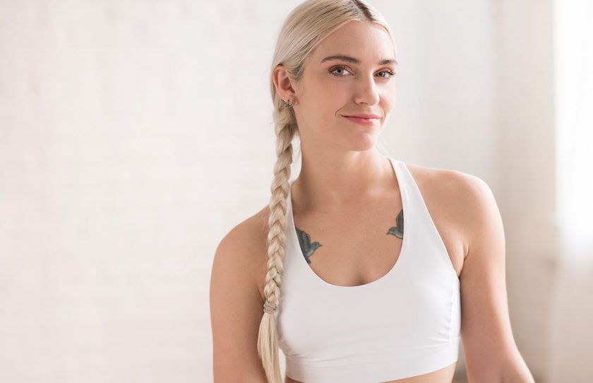 A woman with long blonde hair in a braid, wearing a white sports bra, smiles slightly while standing in a bright, softly lit room. She has visible tattoos on her chest—ready for her NEW YEAR’S RESOLUTION RESCUE at NEW YORK PILATES.