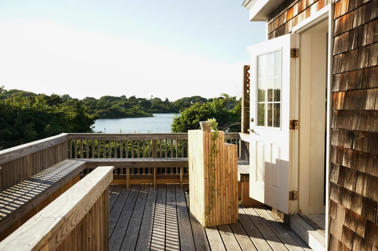 A wooden deck with railing and benches overlooks a scenic view of trees and a lake under a clear sky. An open door leads from the New York Pilates Opens Montauk Studio onto the sunlit deck, where a small table with a plant is placed.
