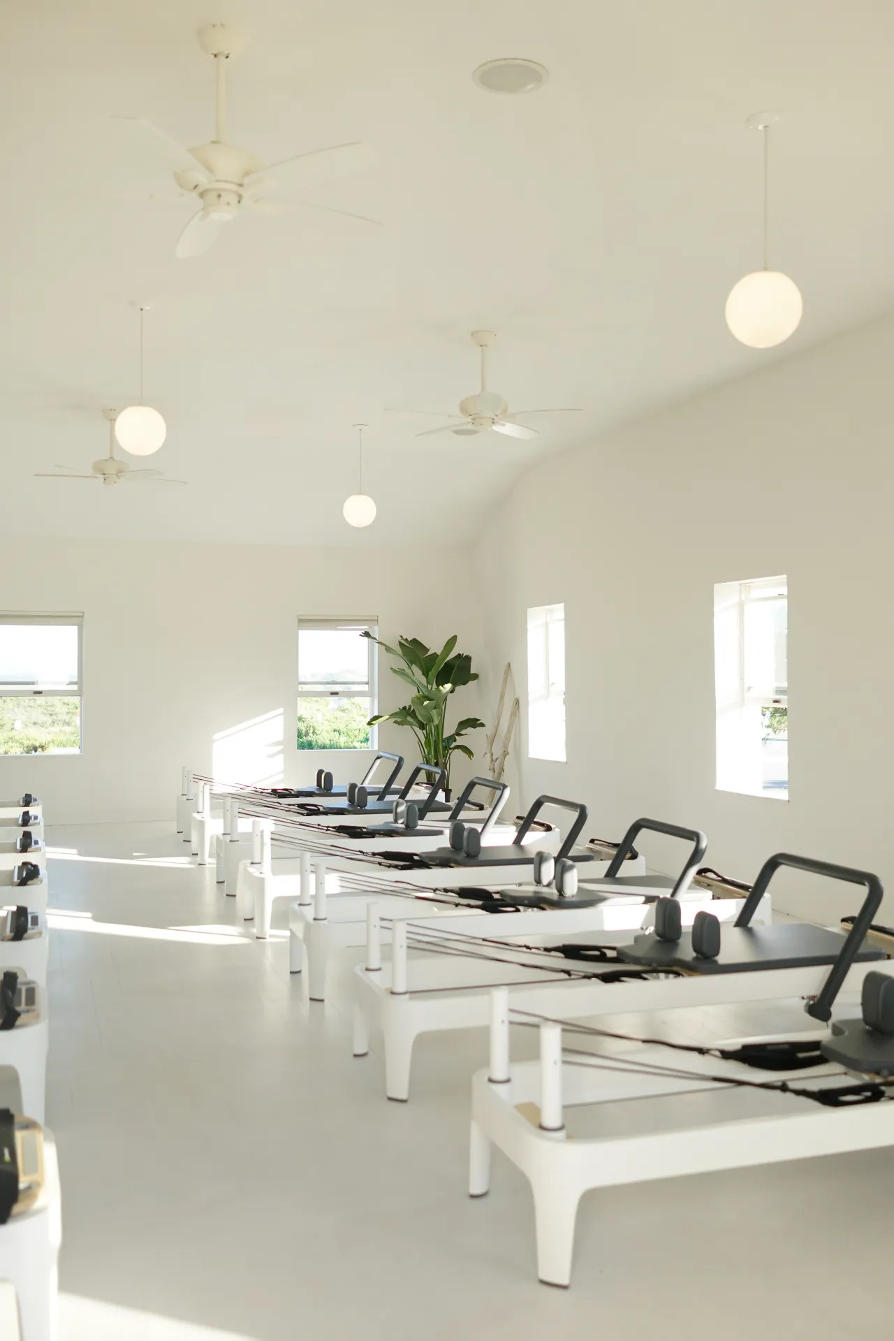 A bright, minimalist Pilates studio welcomes you as New York Pilates opens Montauk Studio—featuring rows of white reformer machines, light streaming through large windows, white walls, ceiling fans, and a potted plant in the corner.