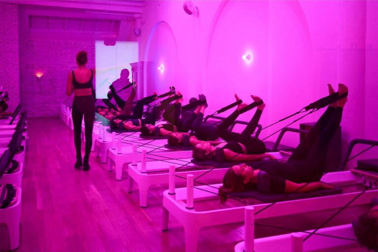 A group of people exercise on Pilates reformer machines in a studio bathed in pink lighting, while an instructor stands at the front guiding the class.