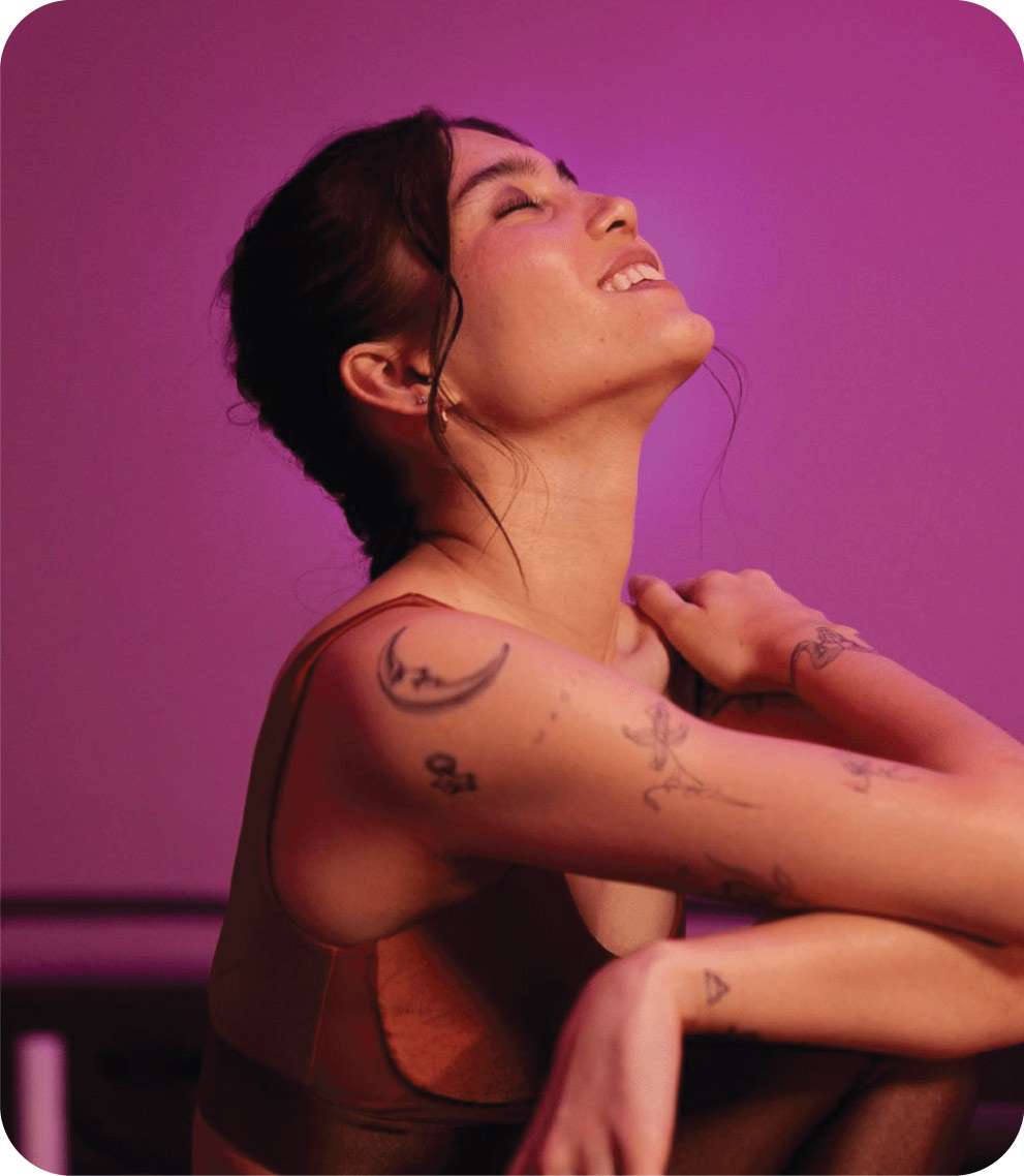 A woman with tattoos on her arm leans back with her eyes closed and smiles, bathed in soft pink and purple lighting, embodying the carefree vibe of Hamptons Intro Offers.