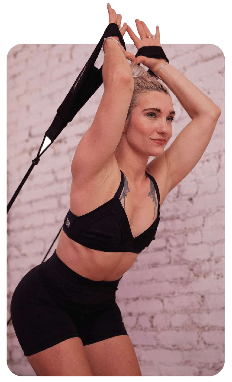 A woman in athletic wear performs a resistance band exercise, arms raised and bent behind her head, in front of a white brick wall. She looks focused and confident—perfect inspiration for anyone pursuing Pilates Instructor Certification.
