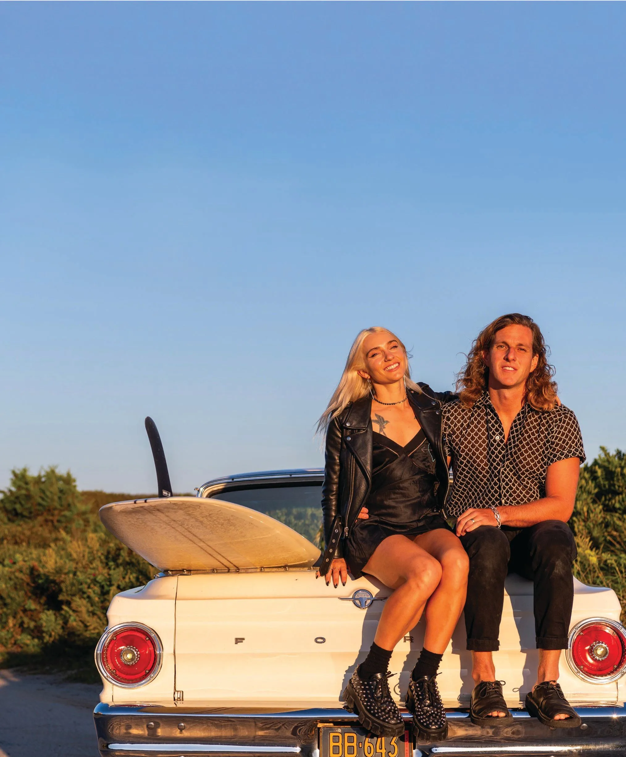 A smiling woman and man, the New York Pilates founders, sit on the trunk of a vintage white car with a surfboard in sunny weather. The woman sports a black outfit, and the man wears a patterned shirt—relaxing amid greenery in the background.