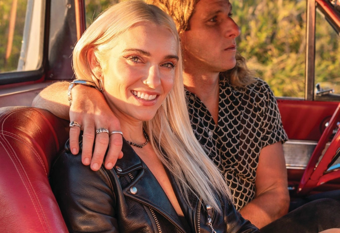 A smiling woman with long blonde hair and a black leather jacket sits in a car, beside one of the New York Pilates founders. Sunlight filters through the window, creating a warm atmosphere that reflects their luxury pilates experience.