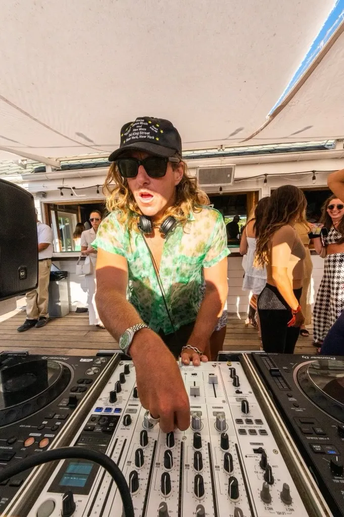 Brion Isaacs in sunglasses, a black hat, and a green tie-dye shirt adjusts controls on a mixing board at an outdoor event for Heather Andersen New York Pilates, while people socialize in the background.