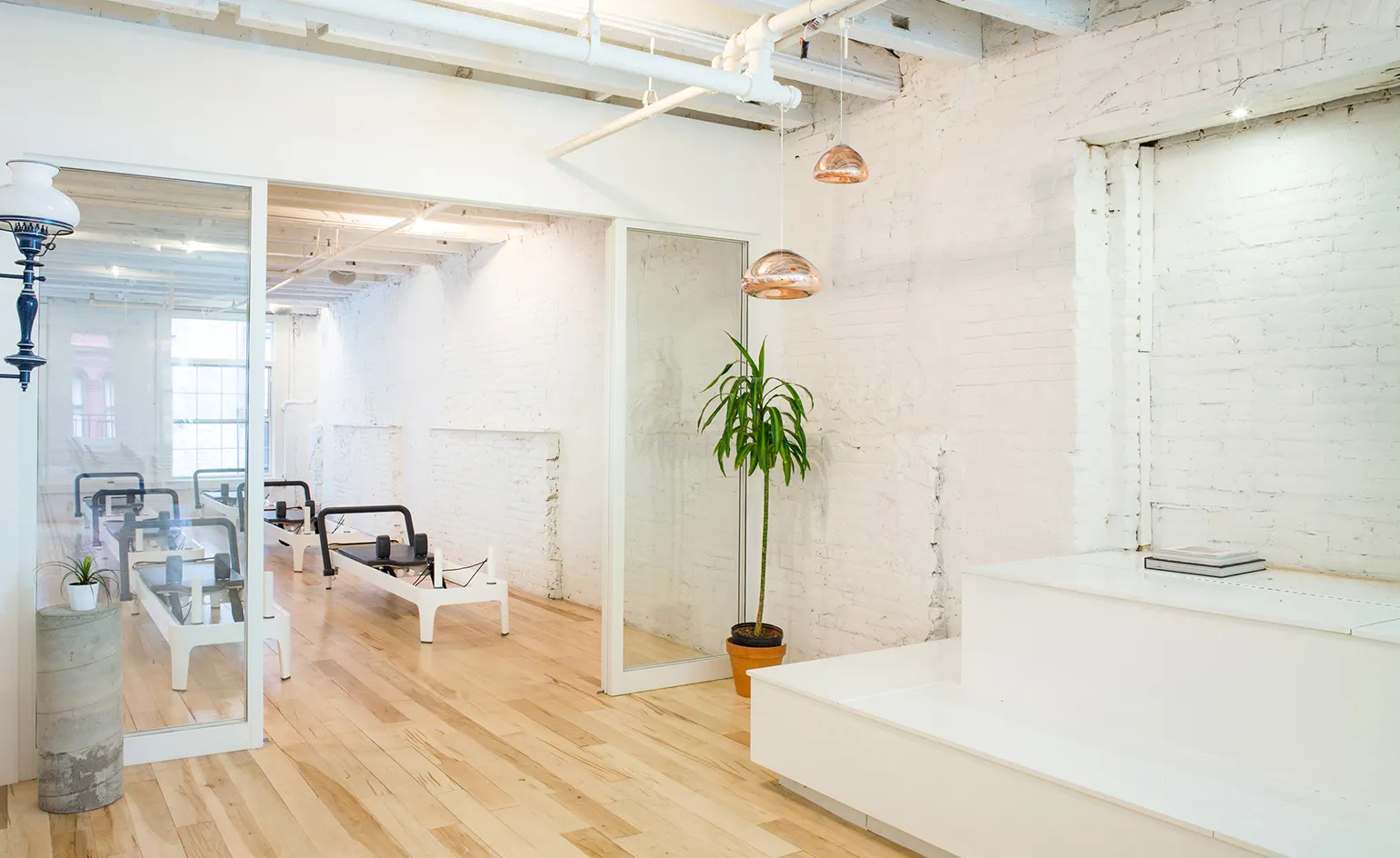A bright, modern Pilates studio inspired by New York Pilates Hamptons, featuring white brick walls, wooden floors, glass partitions, Montauk reformer Pilates machines, a potted plant, and white tiered seating with a closed laptop on top.