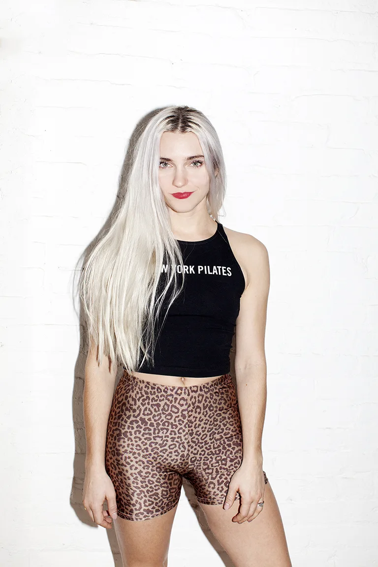 Heather Andersen New York Pilates Founder with long, platinum blonde hair stands against a white brick wall, wearing a black sleeveless crop top that says NEW YORK PILATES—founded by the New York Pilates founders—and high-waisted leopard print shorts. She looks at the camera and smiles slightly.