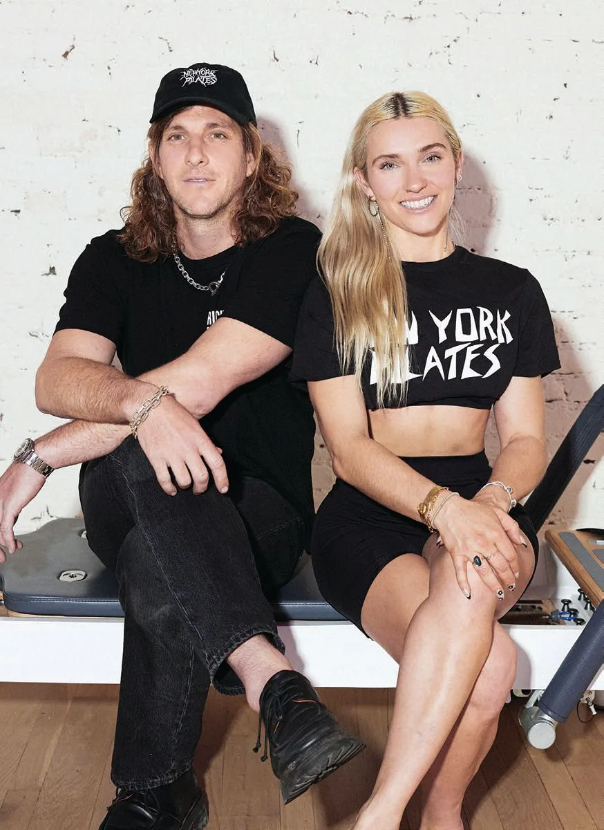 Brion isaacs and heather Andersen sit side by side on exercise equipment. The woman, one of the Pilates founders, wears a black New York Pilates crop top and shorts. Both are smiling against a white brick wall, New York Pilates Founders' vibe.