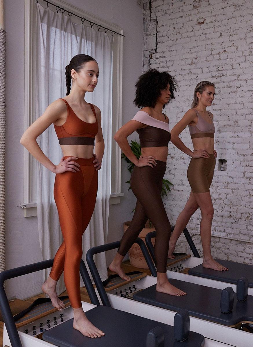 Three women in matching sports bras and earth-tone leggings stand on Pilates reformer machines in a bright studio with white brick walls, large windows, and hardwood floors—an inspiring scene reminiscent of the New York Pilates founders.