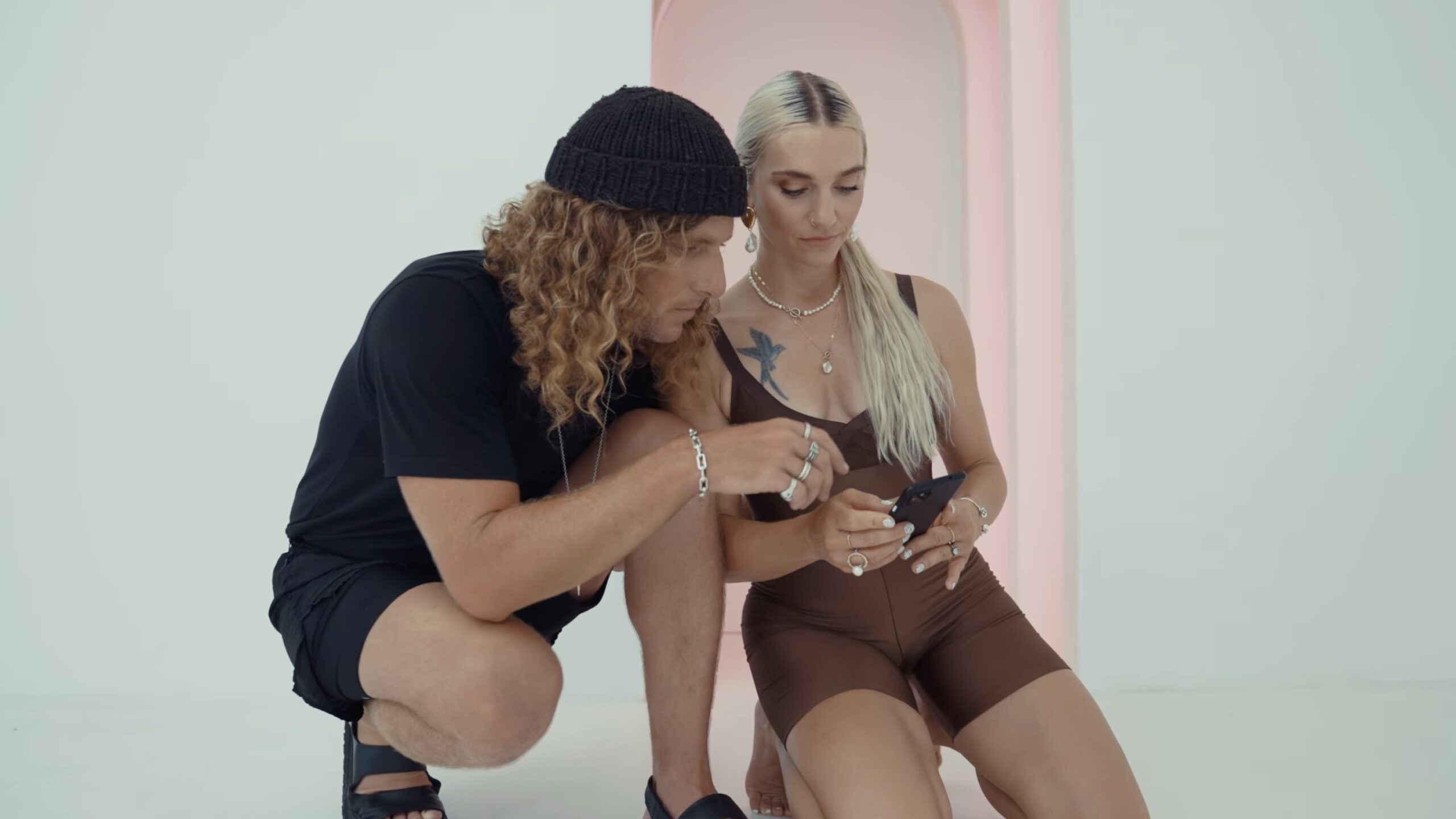 A man in a black outfit and beanie crouches next to a woman in brown with platinum blonde hair. They’re both looking at her phone, possibly searching for a "pilates class near me," in a minimal white space with a pink arch behind them.