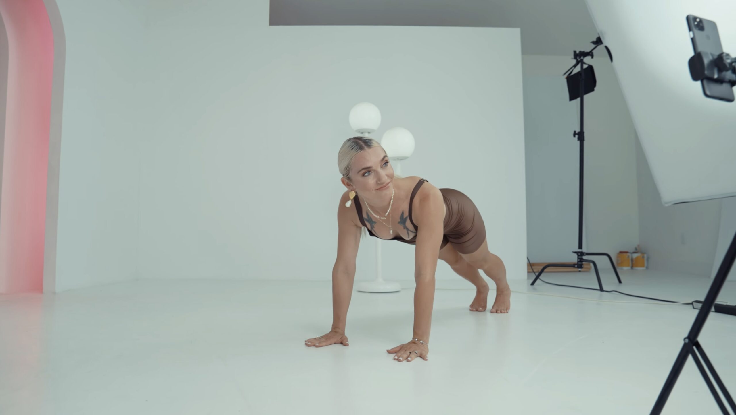 A woman in athletic wear holds a plank position on a white floor in a bright, minimalist studio. She looks to the side, with photo equipment and a lamp visible—capturing the Benefits of Pilates at New York Pilates SoHo.