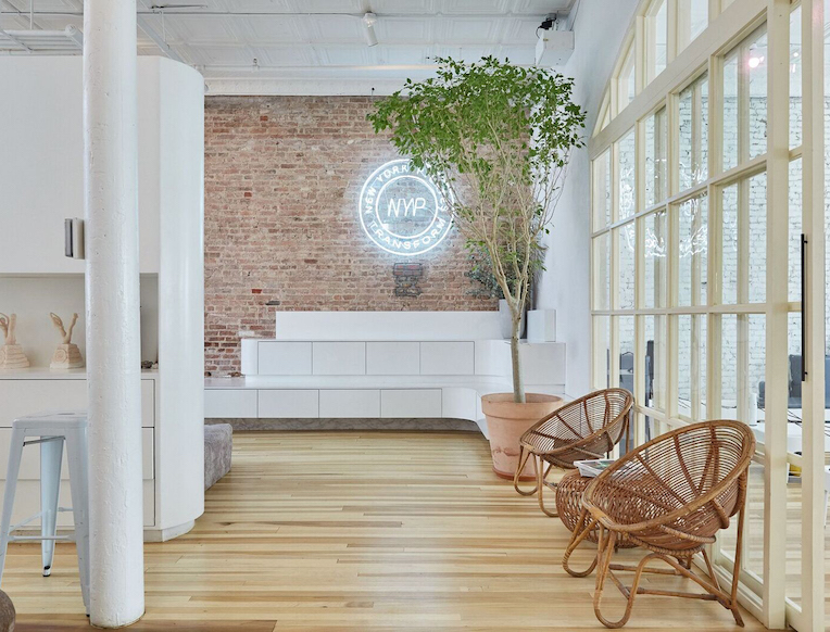 Modern, airy room with a brick wall, white furniture, a large potted tree, rattan chairs, and a neon NYR sign—perfect for a boutique fitness NYC vibe. Light wood floors and large windows create a bright, minimalist atmosphere.