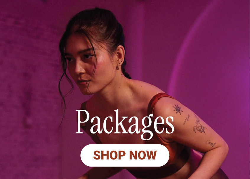 A woman with tattoos on her arm leans forward in a dim, pink-lit room. Text on the image reads Packages and Shop Now in bold white and orange letters, highlighting the Best Pilates in NYC experience.