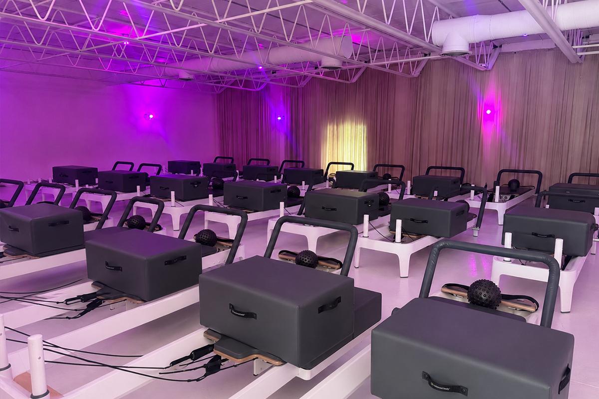 A spacious, modern Sag Harbor Pilates studio with rows of reformer machines neatly arranged under exposed white ceiling beams and purple mood lighting. Light streams through floor-to-ceiling curtains at the back—Reformer Pilates Sag Harbor at its best.