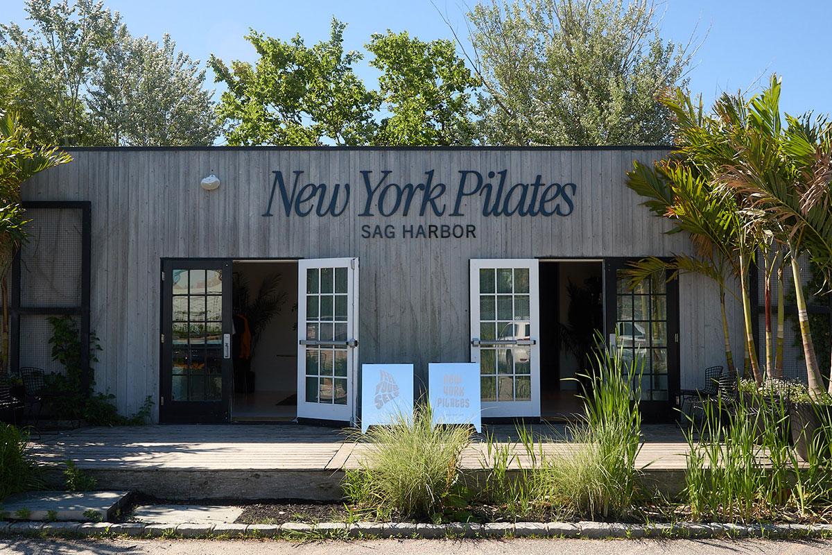 A wood-paneled building with double doors open, displaying New York Pilates Sag Harbor on the front. Lush green plants and trees surround the entrance, hinting at the serene vibe of a top Bridgehampton Pilates Studio just minutes from Reformer Pilates Bridgehampton classes.