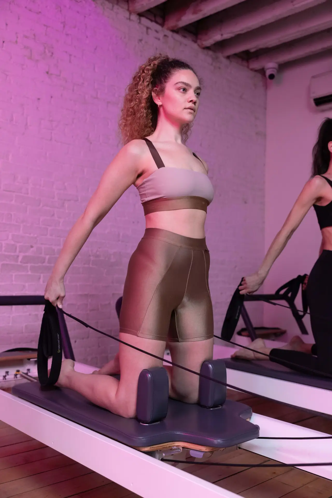 New York Pilates: A woman kneels upright on a reformer while holding resistance straps.