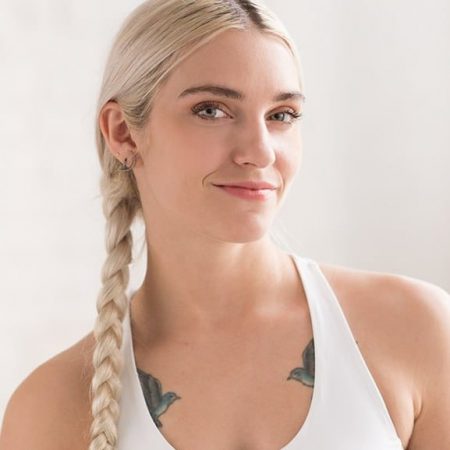 A woman with long, platinum blonde hair in a braid smiles slightly. She wears a white tank top and has bird tattoos on her upper chest—just like those who learned from the team that revitalized reformer Pilates. The background is softly blurred and light.